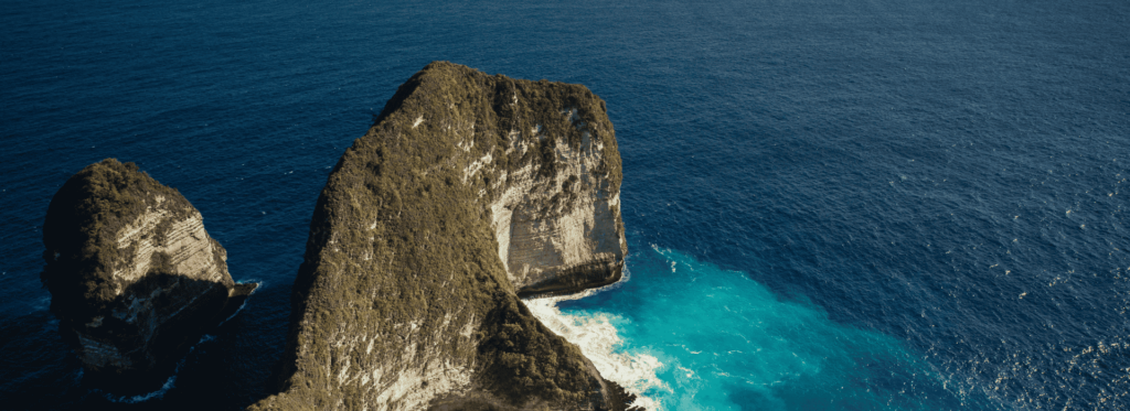 A majestic rock formation rises from the ocean, surrounded by waves and reflecting the sunlight above.