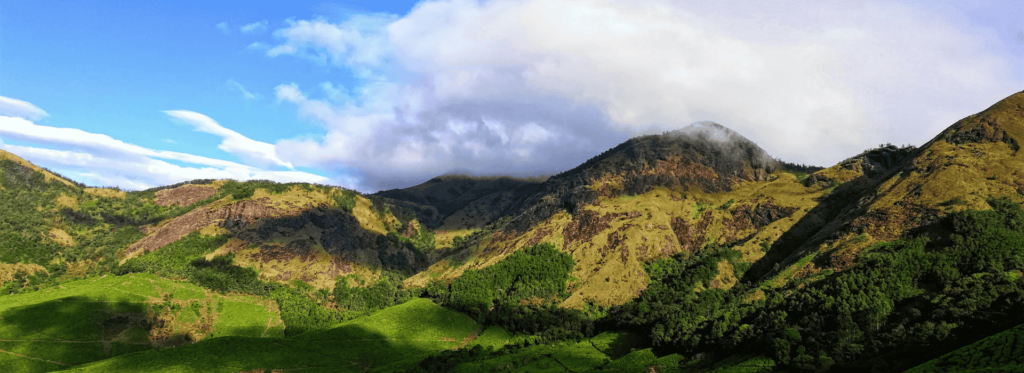 A stunning mountain landscape featuring lush green grass and fluffy clouds in a clear blue sky.